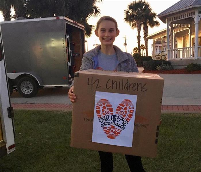 Young girl stands proudly holding a large box full of shoes that were donated.
